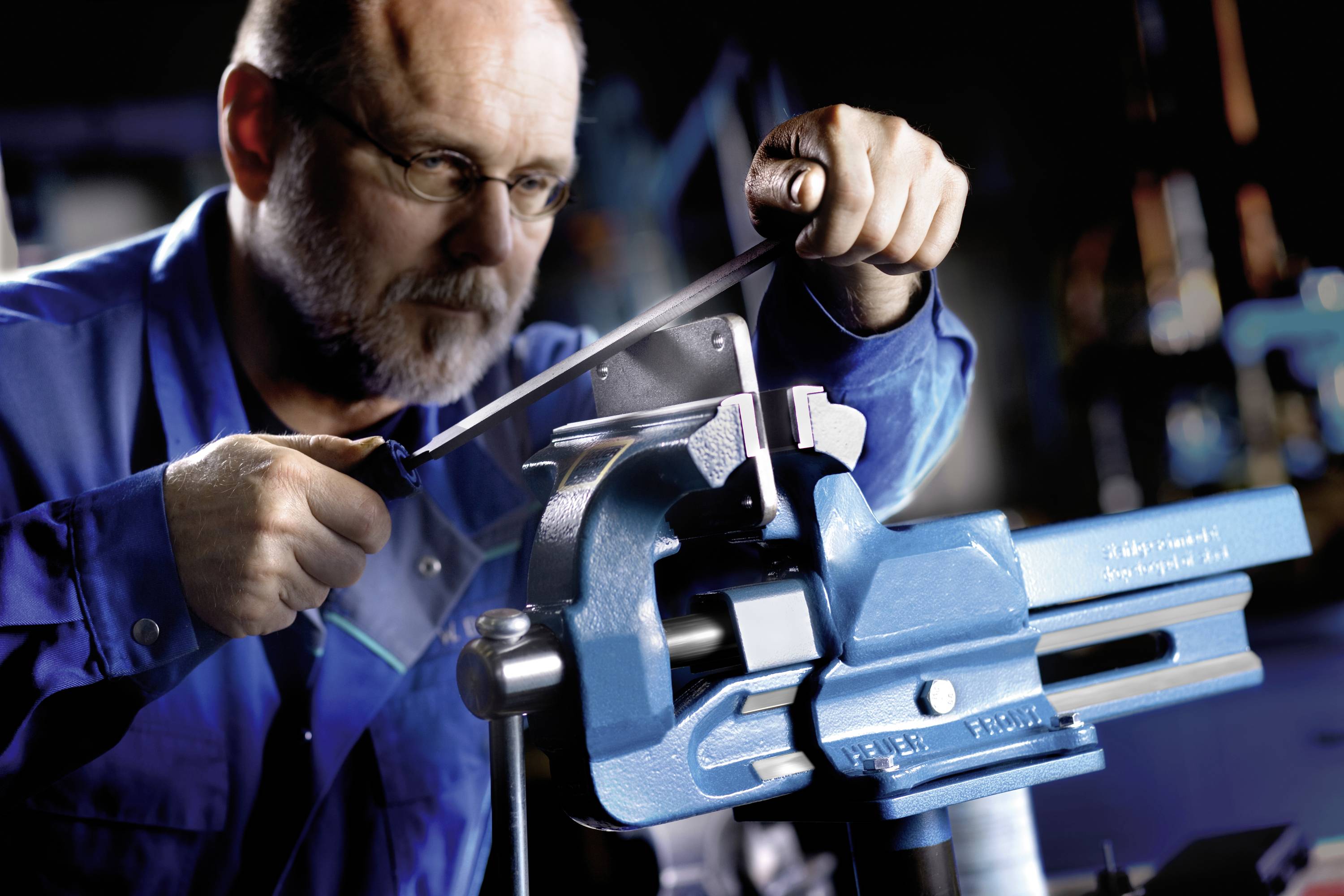 Ein Mann in blauer Arbeitskleidung feilt mit einem Werkzeug an einem Metallstück, das in einem Schraubstock eingespannt ist.