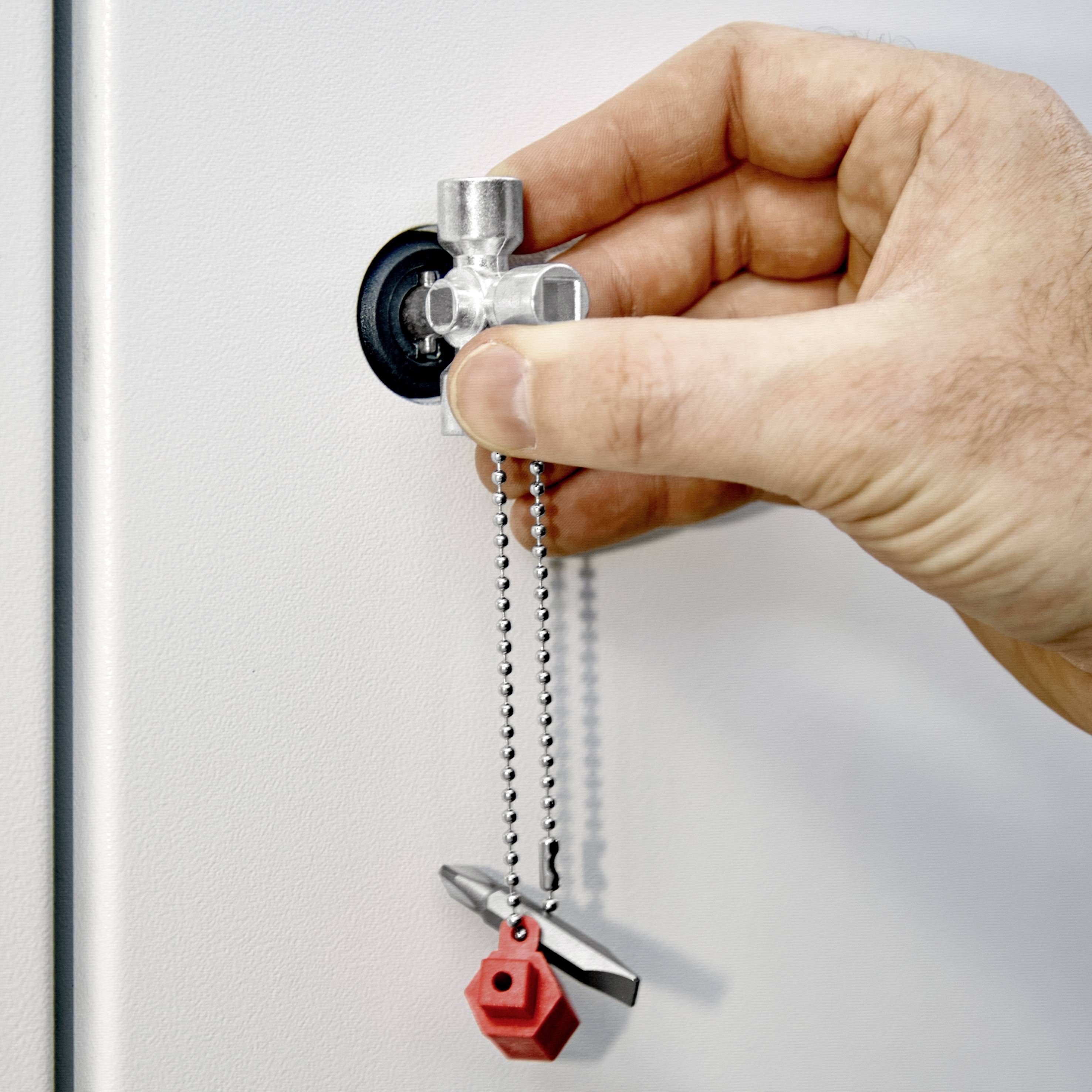 Hand dreht einen Wasserhahn auf, an dem ein Sicherheitsventil mit rotem Schlüsselanhänger und Metallkette hängt, vor grauer Wand.