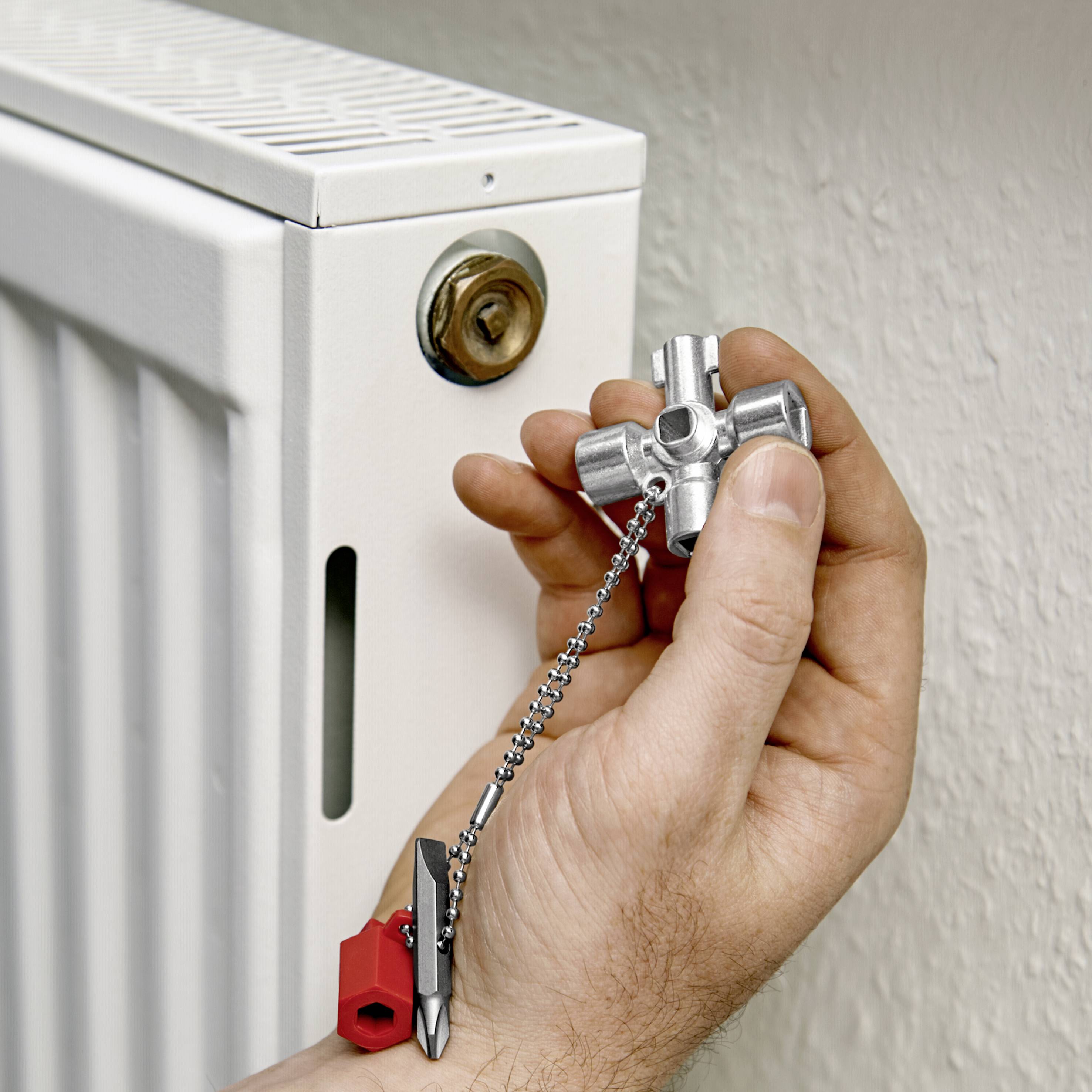 Eine Hand hält einen Entlüftungsschlüssel vor einem weißen Heizkörperventil, bereit zur Entlüftung des Heizkörpers.