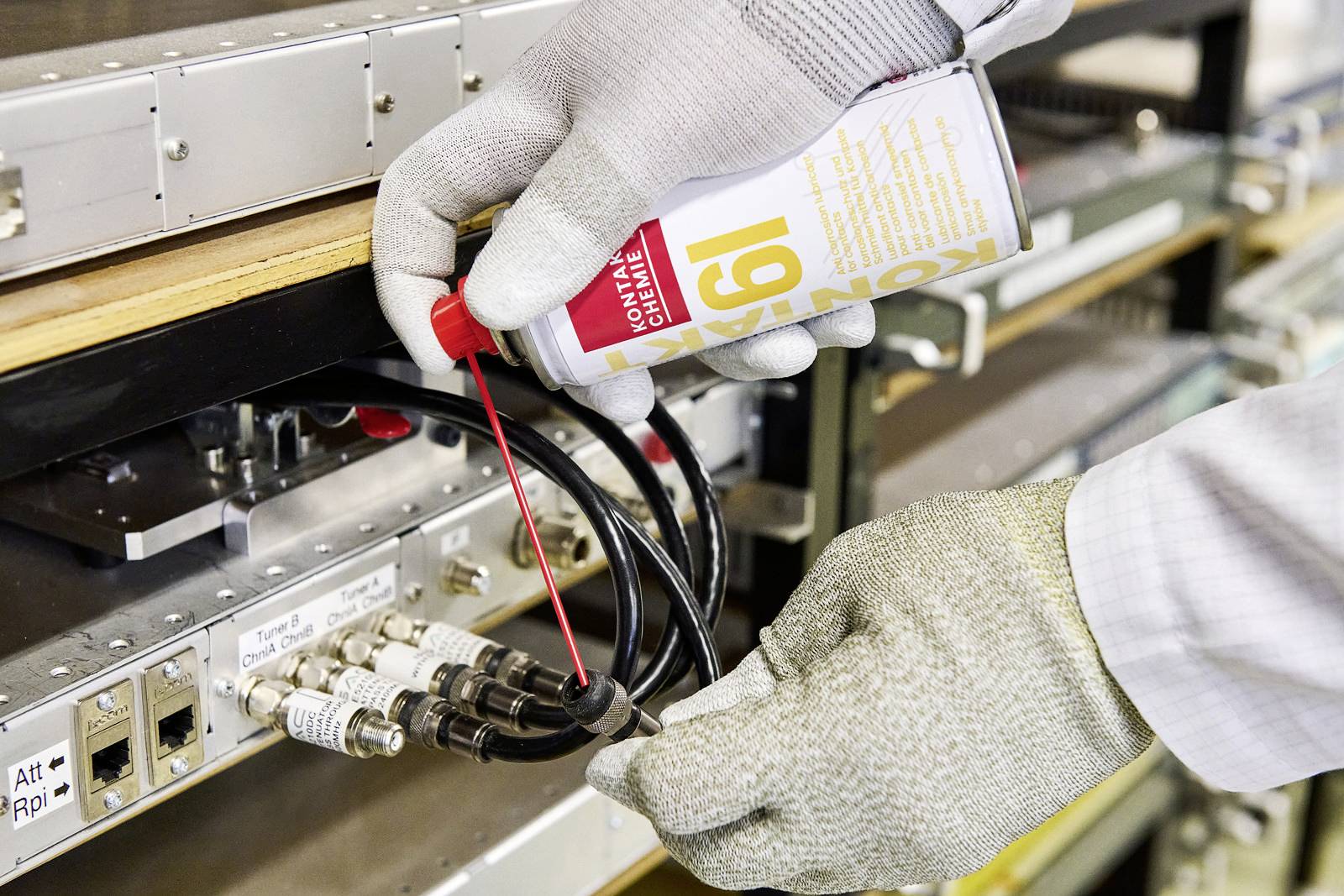 Eine Person mit Handschuhen reinigt Kabelanschlüsse an einer technischen Anlage mit einem Spray.