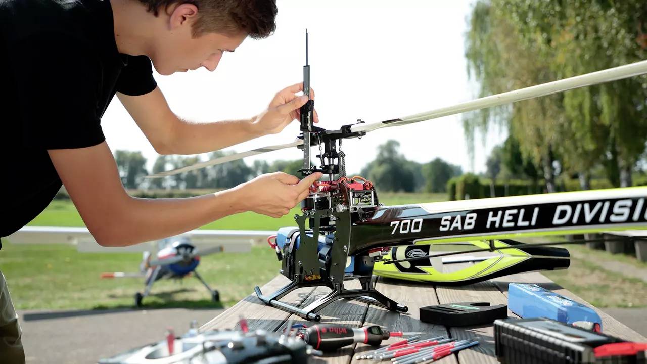 Ein Mann arbeitet im Freien an einem Modellhubschrauber mit der Aufschrift 'SAB Heli Division'. Werkzeug und Zubehör sind auf dem Tisch.