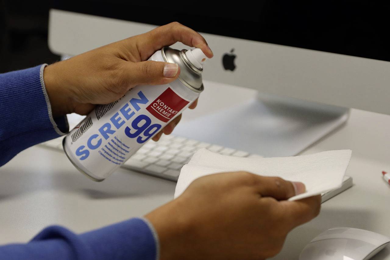 Eine Person sprüht Bildschirmreiniger 'SCREEN 99' auf ein Tuch vor einem Computer, um die Reinigung vorzubereiten.