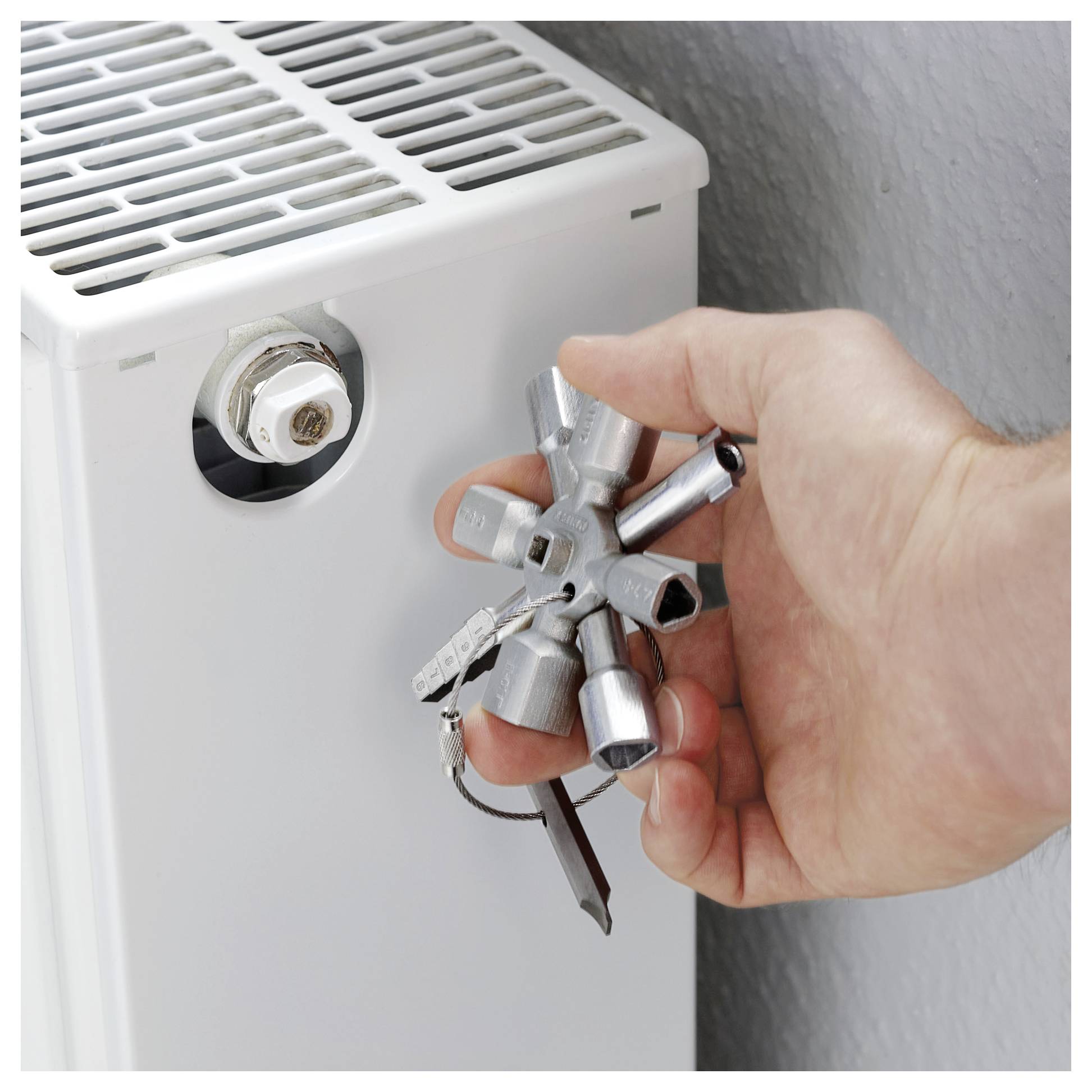 Hand, die ein Multitool verwendet, um einen Heizkörper zu entlüften, während sie das Ventil an einem weißen Heizkörper mit einer Wand im Hintergrund verstellt.