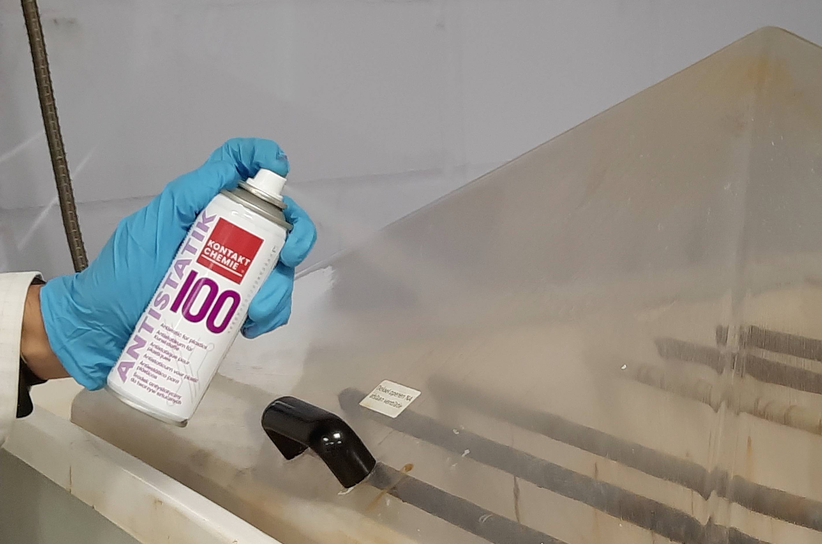 Jemand mit blauen Handschuhen hält eine Dose Antistatik-Spray vor einer Kunststoffabdeckung in einer Werkstatt oder Laborumgebung.