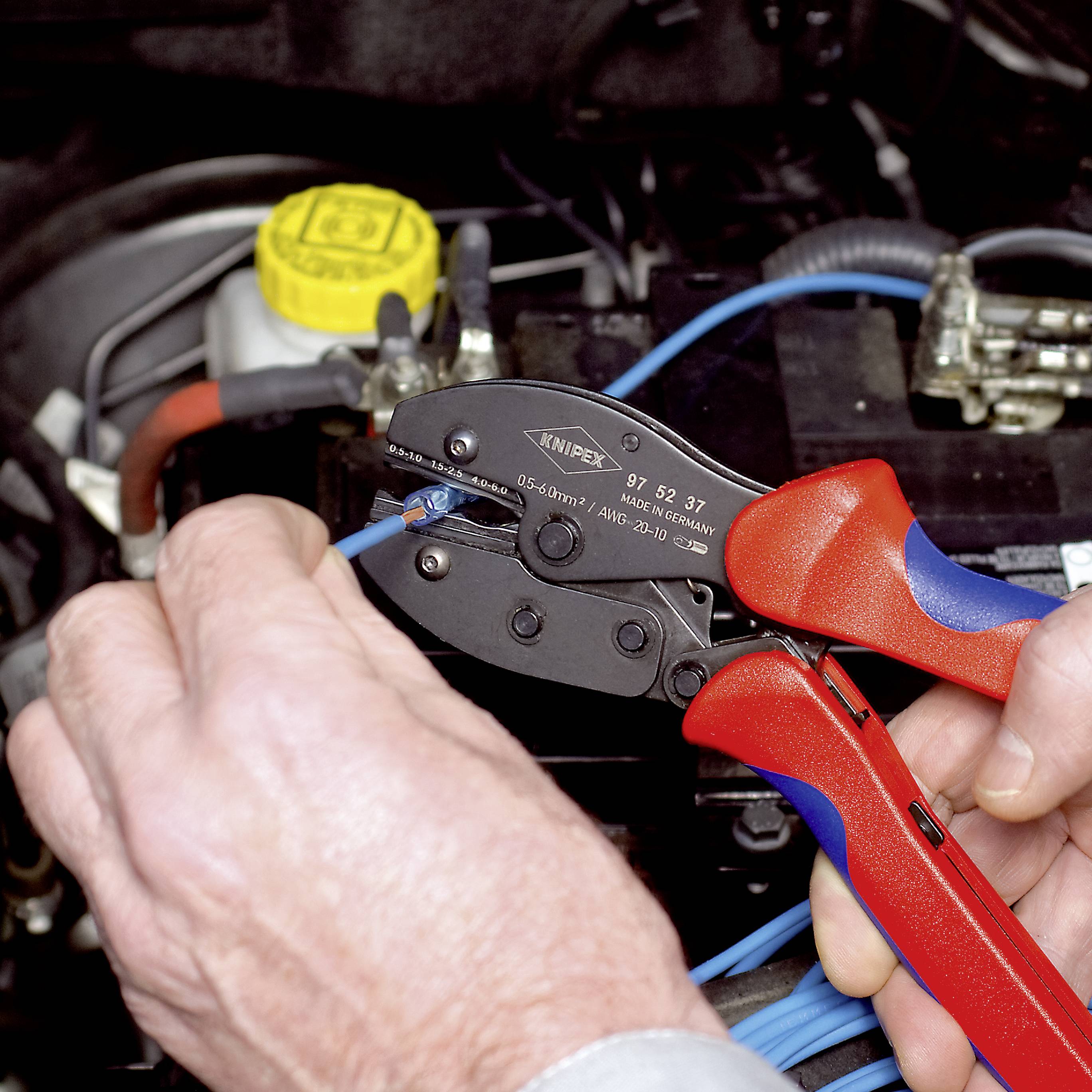 Person verwendet eine rote und blaue Zange, um eine elektrische Verkabelung an einem Auto zu crimpen. Im Hintergrund ist ein Motorraum zu sehen.