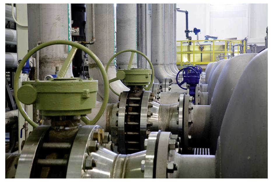 Rohre und Absperrventile in einer Industrieanlage, mit grünen und blauen Ventilrädern und großen Metallrohren vor einem hellen Hintergrund.