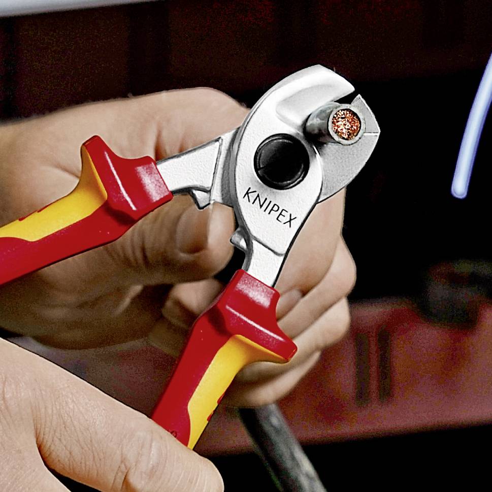 Eine Hand hält einen KNIPEX-Kabelschneider, der ein dickes, kupfernes Kabel durchtrennt. Der Hintergrund ist unscharf.