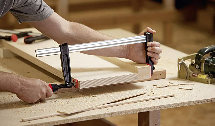 Eine Person befestigt ein Holzstück mit einer Schraubzwinge an einem Tisch. Werkzeuge und Holzspäne sind auf der Werkbank verteilt.