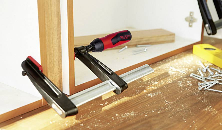 Eine Zwinge hält zwei Holzplatten zusammen, mit Schrauben und Holzspänen auf einem Tisch im Hintergrund, was auf eine handwerkliche Arbeit hinweist.