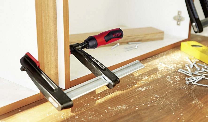Ein Schraubzwinge fixiert zwei Holzstücke an einem Tisch, umgeben von Schrauben und einem gelben Winkelmesser.