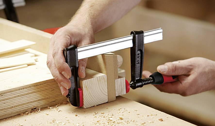 Eine Person verwendet eine Klemme, um zwei Holzstücke auf einer Werkbank zusammenzuhalten. Holzspäne liegen verstreut auf der Oberfläche.