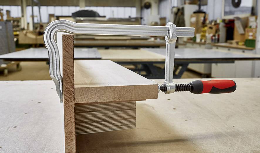 Ein Schraubzwinge hält zwei Holzbretter zusammen in einer Werkstattumgebung. Im Hintergrund sind Werkzeuge und Holzmöbel sichtbar.