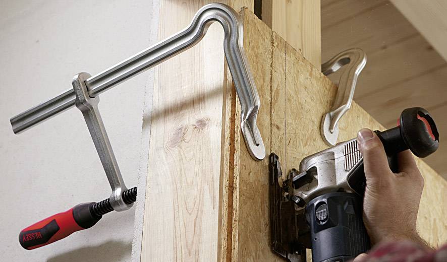 Eine Person bearbeitet mit einer Oberfräse die Kante eines Holzteils, das mit zwei Schraubzwingen an einer Wand befestigt ist.