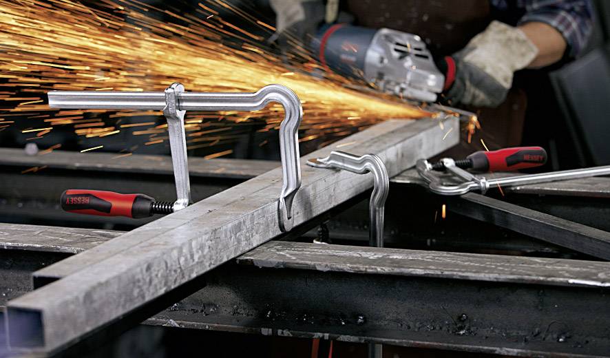 Eine Person schleift ein Metallrohr mit einer elektrischen Schleifmaschine. Funken fliegen vom Kontaktpunkt. Metallschellen fixieren das Rohr.