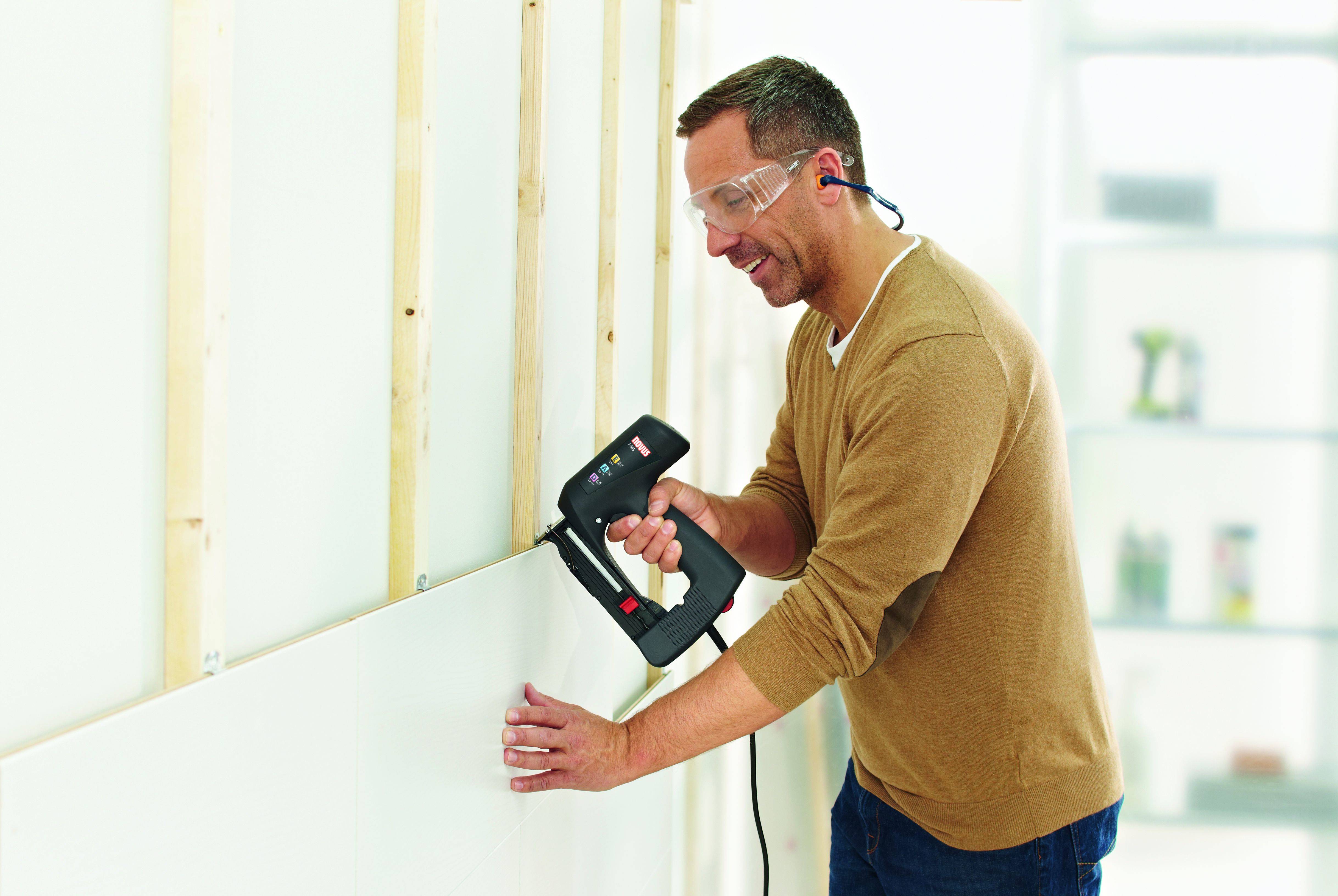 Ein Mann befestigt mit einer Nagelpistole eine Gipsplatte an einer Wand. Er trägt Schutzbrille und Gehörschutz und arbeitet konzentriert.