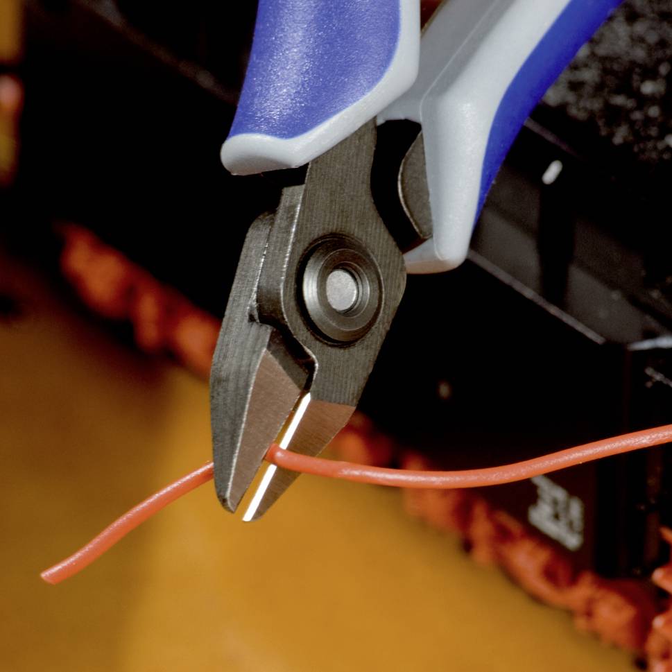 Nahaufnahme einer Zange, die ein rotes Kabel schneidet. Klarer Fokus auf der Schneideaktion, im Hintergrund sind unscharfe Werkbankteile zu sehen.
