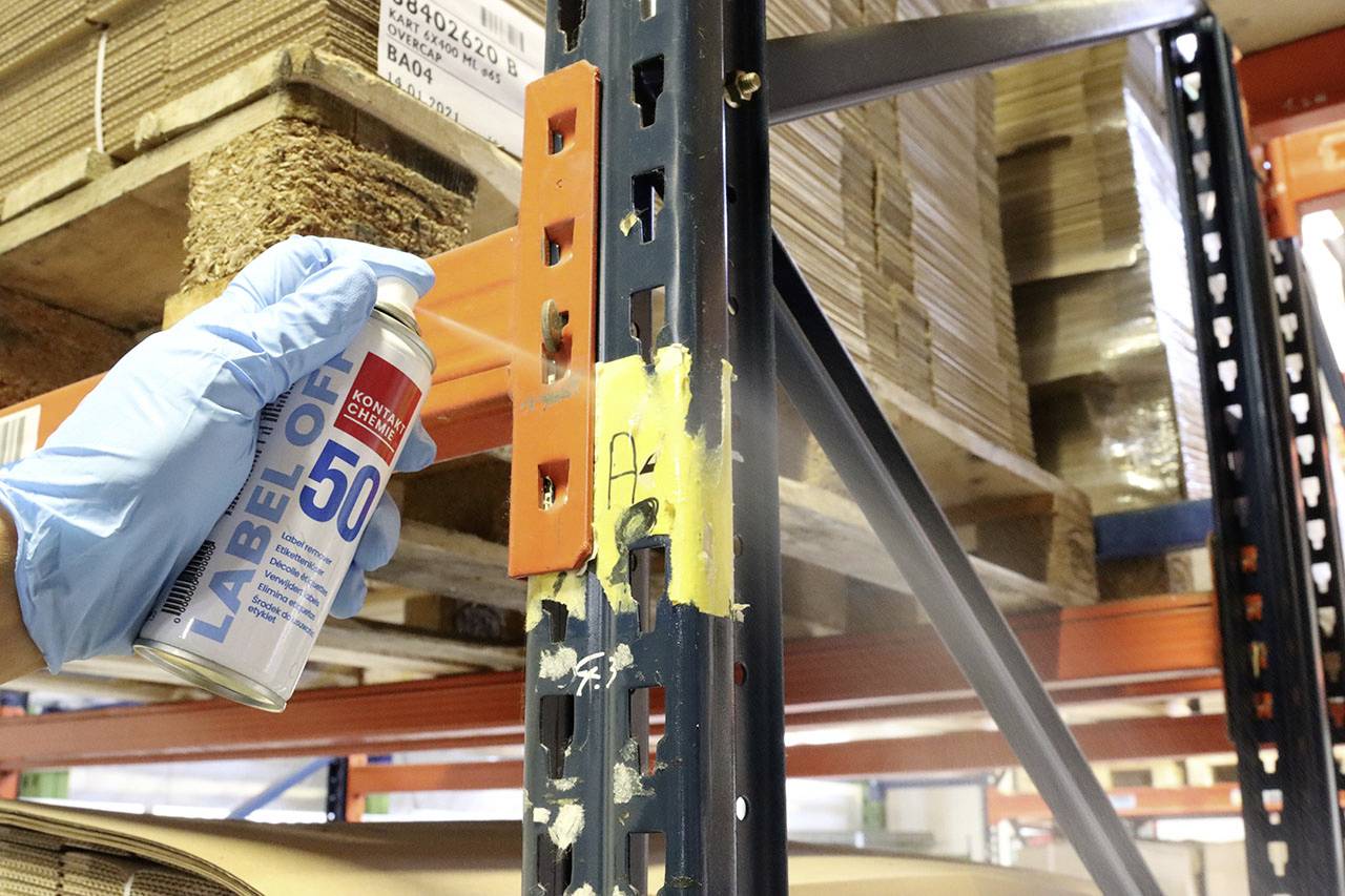 Ein Behandschuhter hält eine Dose Etikettenentferner bei einem Regal in einem Lager, um Etikettenreste von der Metallsäule zu entfernen.