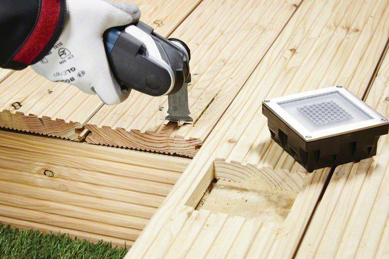 Eine Hand mit Handschuh hält ein elektrisches Werkzeug und schneidet Holzplanken. Auf dem Boden ist eine quadratische Lampe zu sehen.