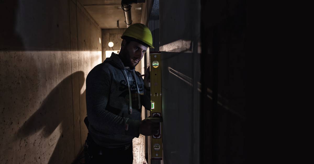 Ein Bauarbeiter mit gelbem Helm benutzt eine Wasserwaage, um eine Wand in einem schlecht beleuchteten Tunnel zu überprüfen.