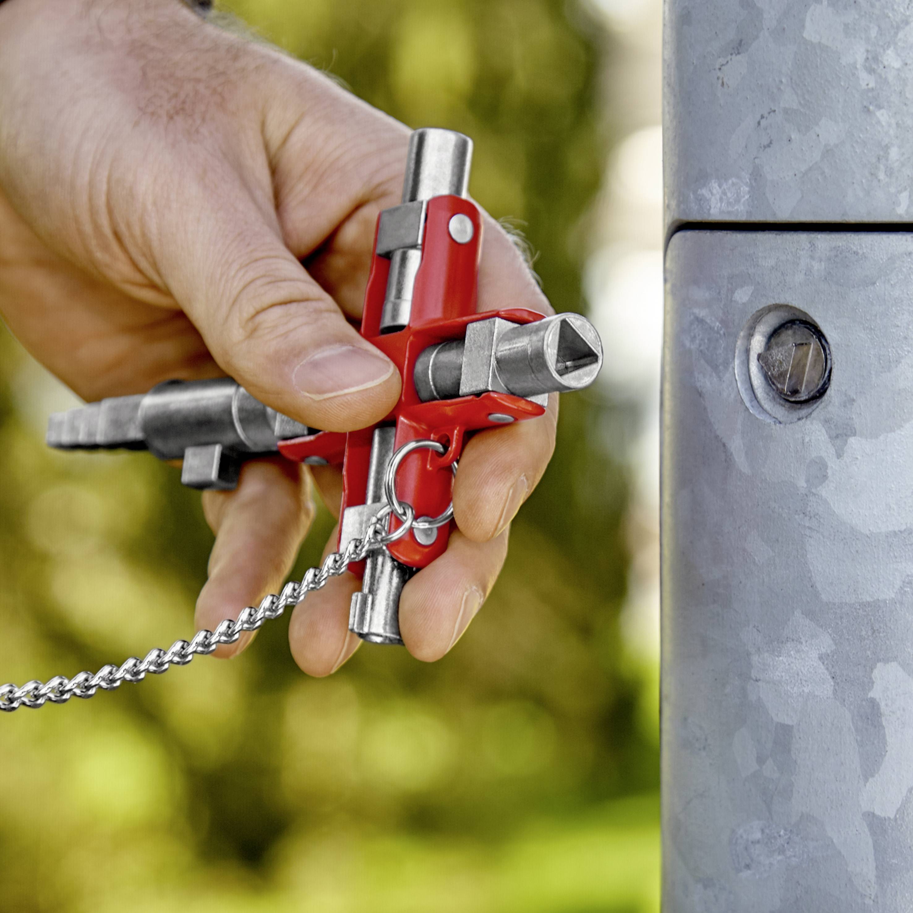 Hand hält einen Metall-Verriegelungsmechanismus an einem Pfosten, gesichert mit einer Kette. Hintergrund ist unscharf.