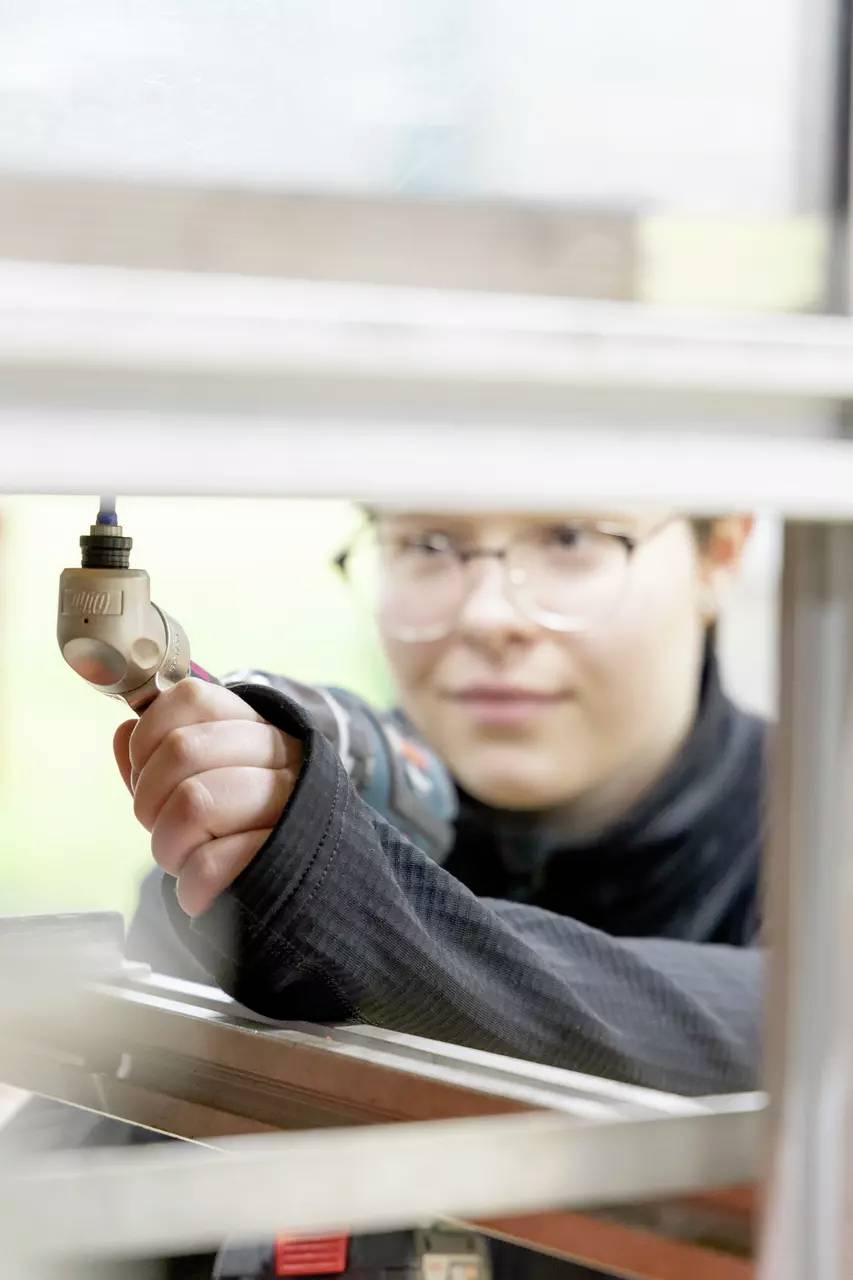 Eine Person mit Brille und kurzem Haar benutzt einen elektrischen Bohrer und konzentriert sich intensiv auf ihre Arbeit durch eine Lücke im Fensterrahmen.