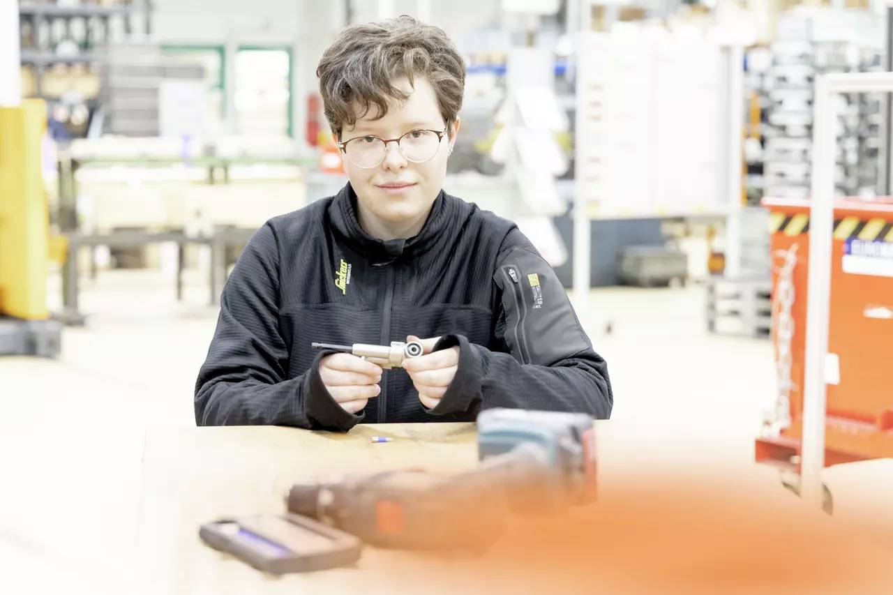 Eine Person mit Brille und einer schwarzen Jacke sitzt an einem Tisch und hält ein kleines Werkzeug in einer hell beleuchteten Werkstattumgebung.