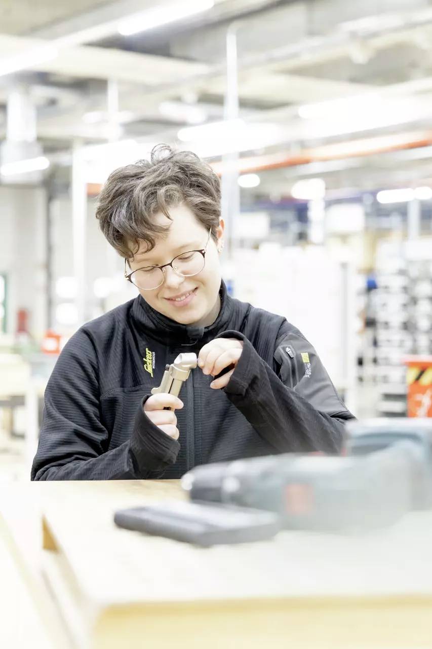Eine Person mit Brille benutzt ein Werkzeug in einer Werkstatt und konzentriert sich auf ihre Aufgabe. Im Hintergrund befinden sich industrielle Ausrüstungen und Regale.