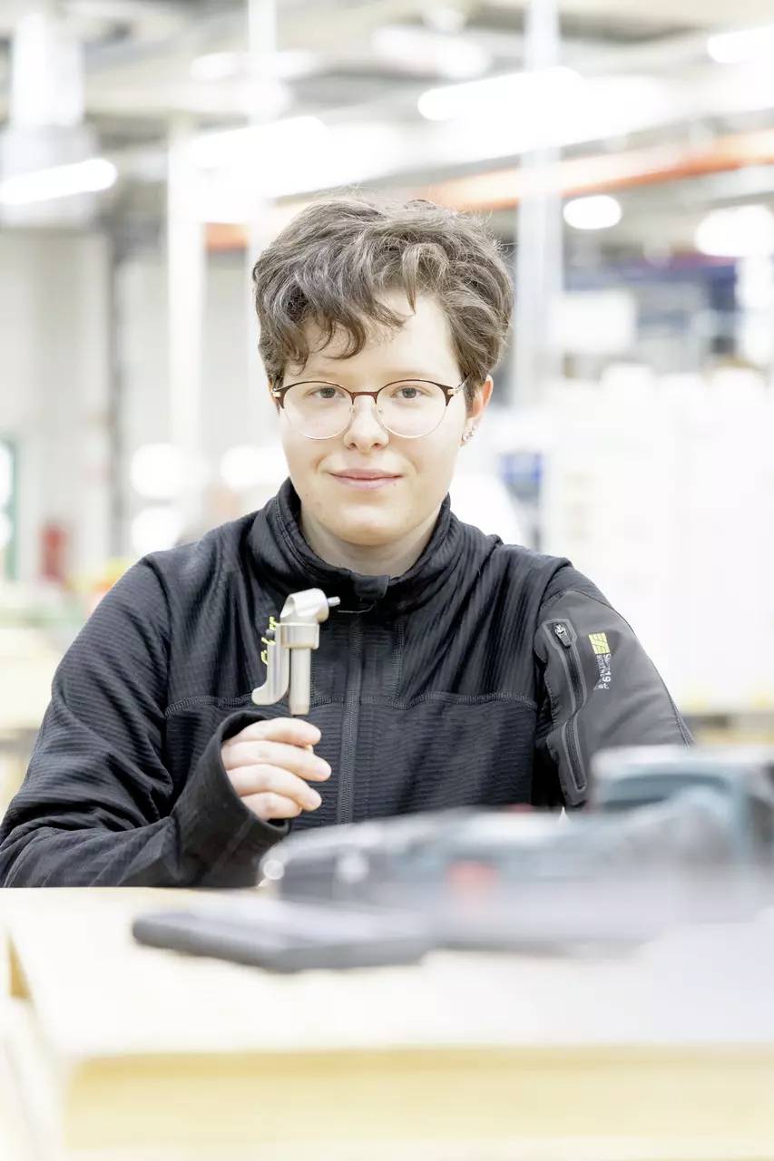 Person, der einen Messschieber in einer Werkstatt hält, trägt eine Brille und eine schwarze Jacke, mit unscharfen Werkzeugen auf dem Tisch im Vordergrund.