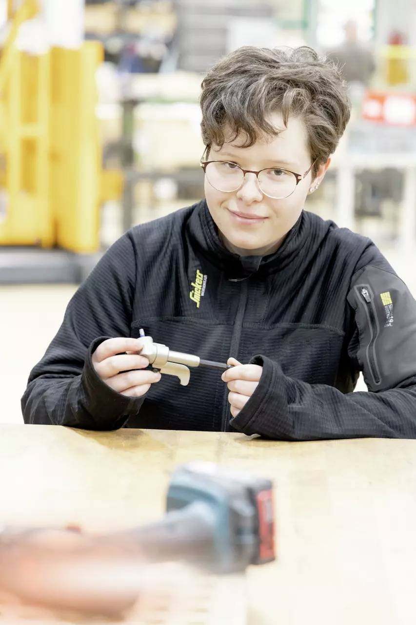 Eine Person mit kurzen Haaren und Brille sitzt an einem Tisch, hält ein kleines Metallwerkzeug und einen Bolzen und trägt eine schwarze Jacke.