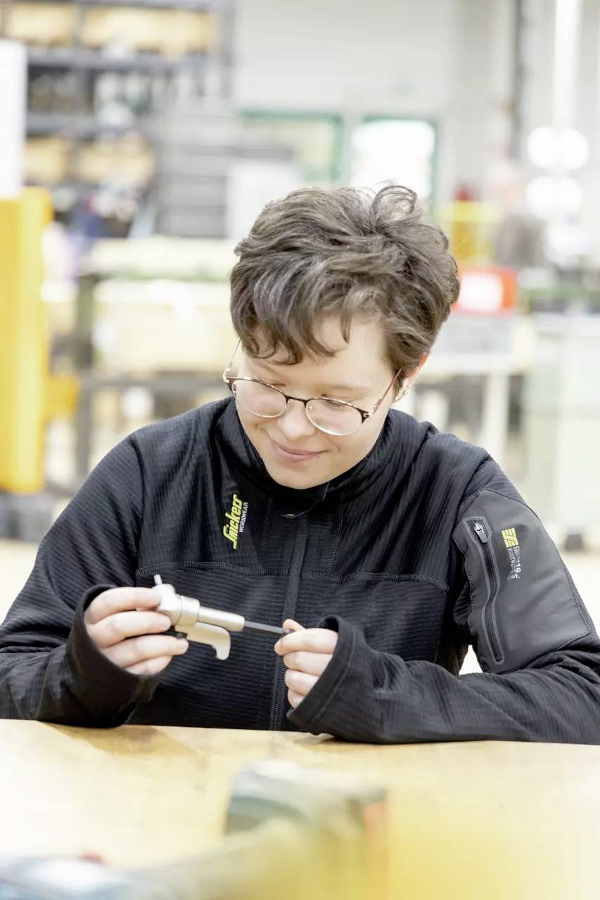 Eine Person mit kurzen Haaren und Brille untersucht an einem Arbeitsplatz ein Metallbauteil und konzentriert sich dabei intensiv auf den Gegenstand. Regale und Ausrüstung sind im Hintergrund unscharf.