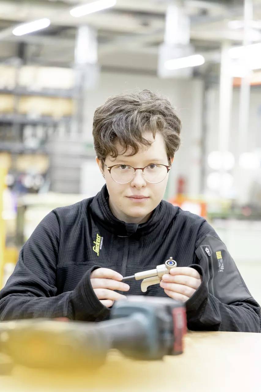 Eine Person mit Brille und schwarzer Jacke untersucht ein kleines mechanisches Bauteil in einer industriellen Werkstattumgebung.