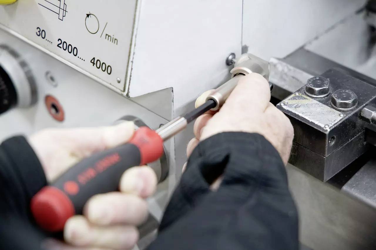 Hände, die einen Schraubendreher verwenden, um ein Maschinenteil einzustellen, neben einem Kontrollpanel mit Geschwindigkeitseinstellungen von 30 bis 2000 Einheiten/min.