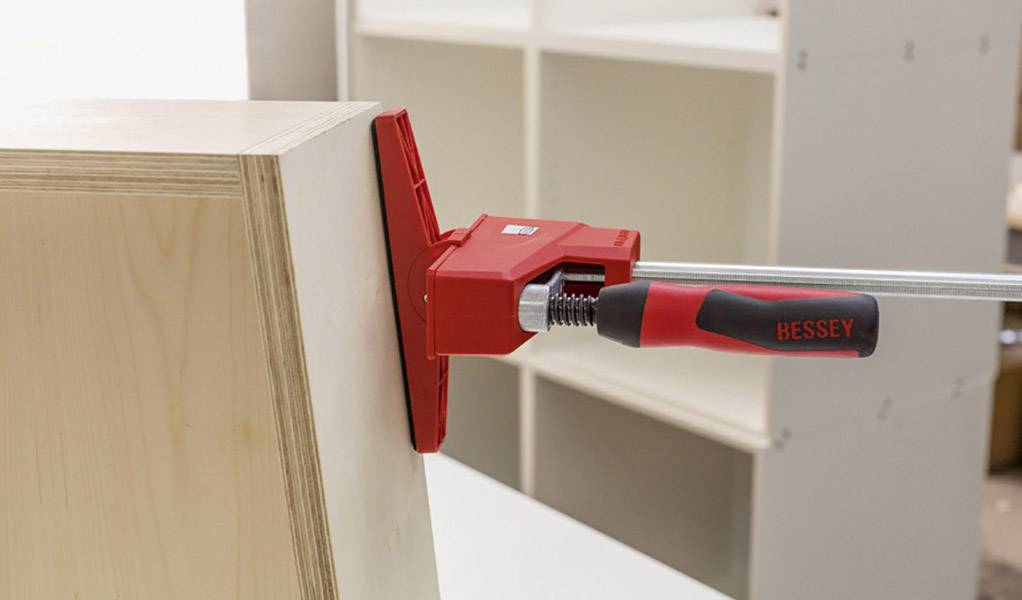Ein Spannsystem mit rotem Griff hält zwei Holzteile zusammen, möglicherweise für Montage oder Tischlerarbeiten in einer Werkstatt.