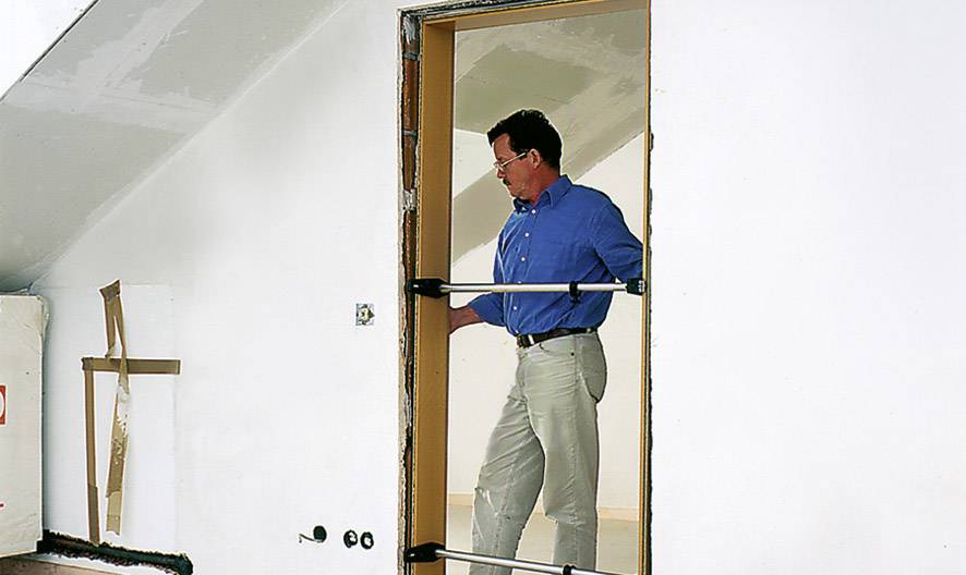 Ein Mann in blauer Arbeitskleidung misst eine Türöffnung in einem weißen Raum mit Hilfe einer Stange zur Vermessung.