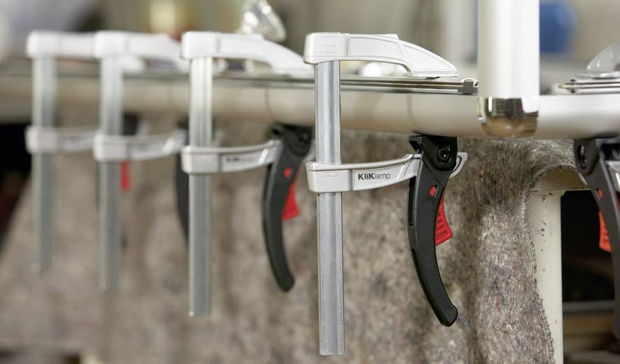 Mehrere Klemmen, die ein Werkstück auf einer Werkbank fixieren. Fokus auf die Klemmen in einer Werkstattumgebung.