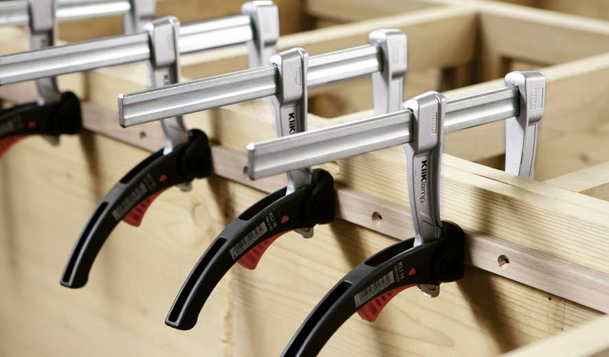 Holzwerkbank mit mehreren festgespannten Schraubzwingen, die ein Brett fixieren.