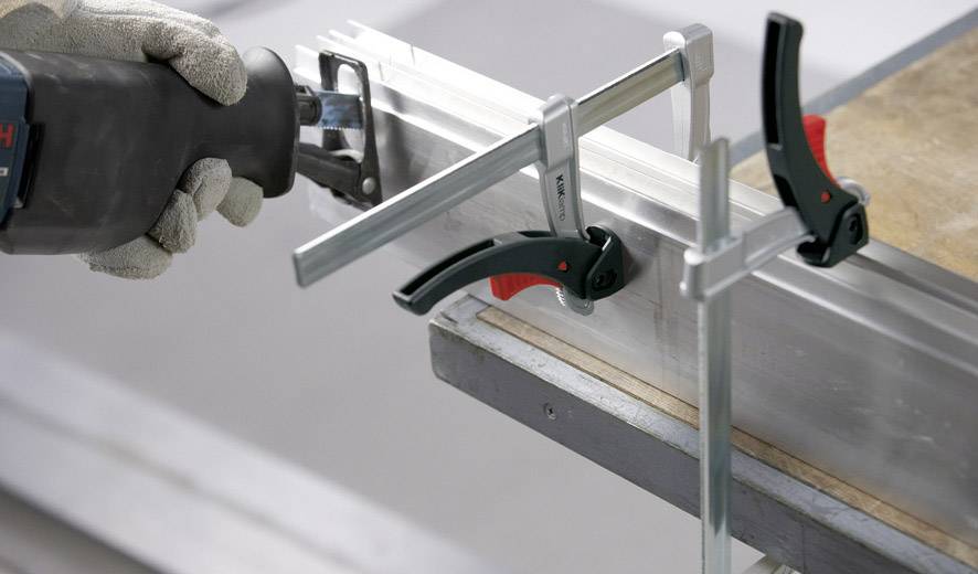 Eine Hand mit Handschuhen hält eine elektrische Säge und schneidet durch ein befestigtes Metallstück, das mit Schraubzwingen auf einem Tisch gesichert ist.
