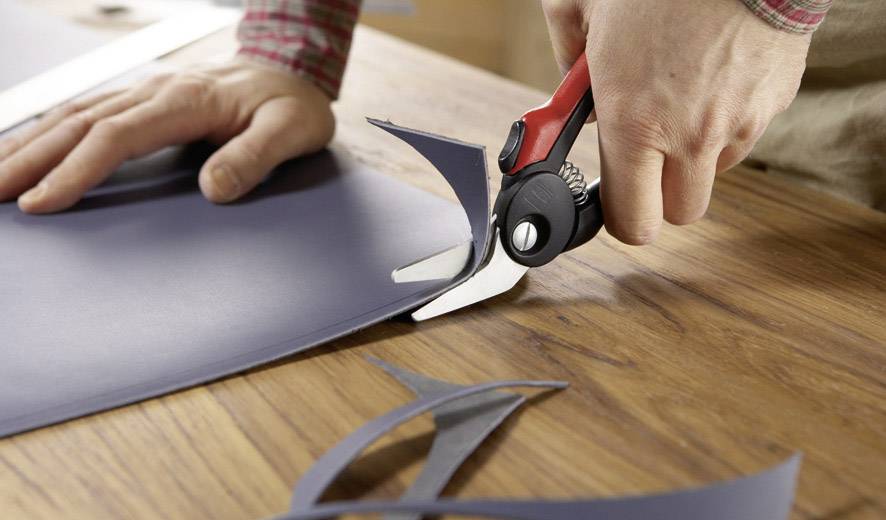 Person schneidet mit einer Metallschere grauen Karton auf einem Holztisch. Konzentration auf präzise Handarbeit.