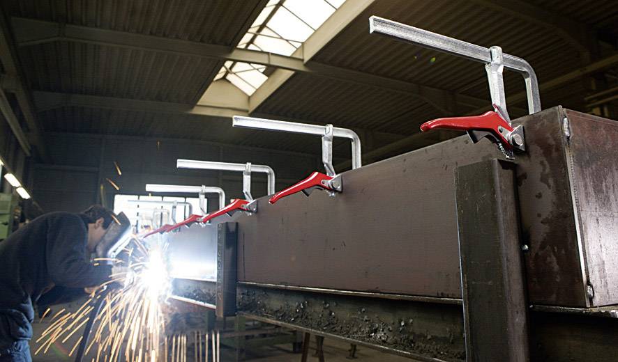 Ein Metallarbeiter schweißt ein großes Metallstück in einer Fabrikhalle. Funken sprühen. Große rote Schraubzwingen halten das Werkstück fest.