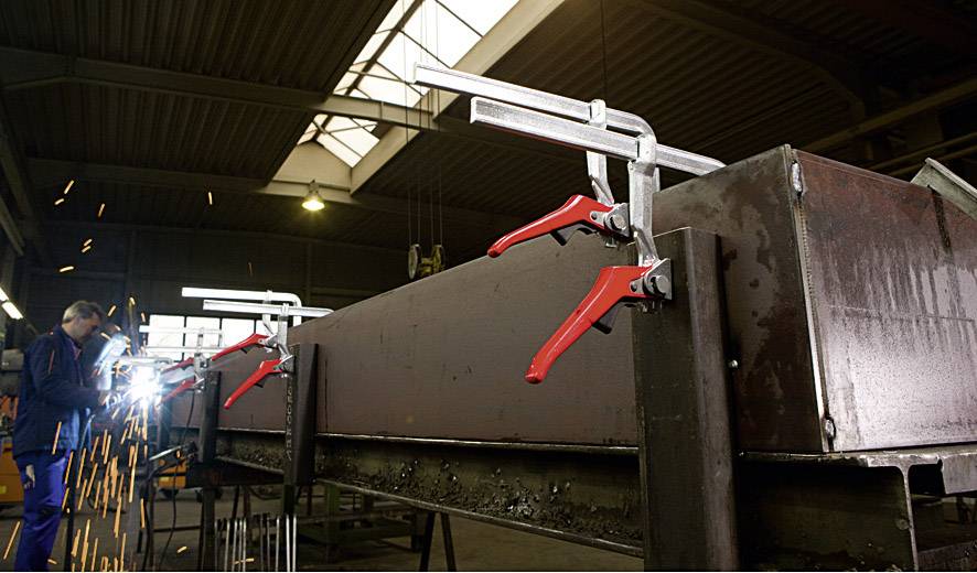 Eine Person schweißt in einer Werkstatt, Funken fliegen durch die Luft. Große Metallzwingen sind an einem Werkstück befestigt.
