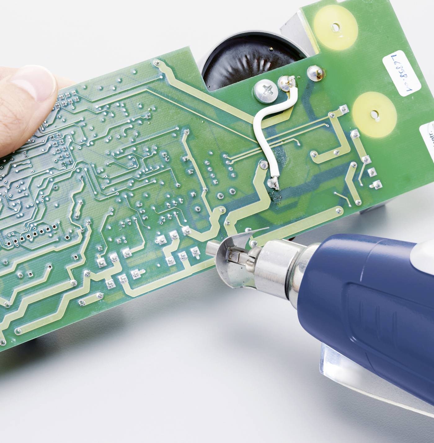 Person hält eine Leiterplatte, während sie mit einem Heißluftgebläse bearbeitet wird. Der Fokus liegt auf der Schaltung.