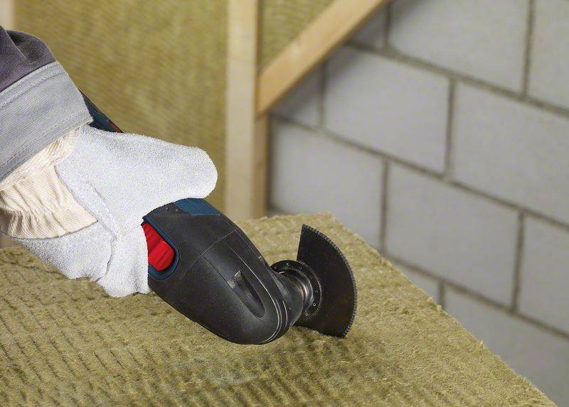 Eine Person schneidet mit einem Elektrowerkzeug eine Dämmplatte. Rechts im Hintergrund ist eine Mauer aus grauen Ziegeln zu sehen.