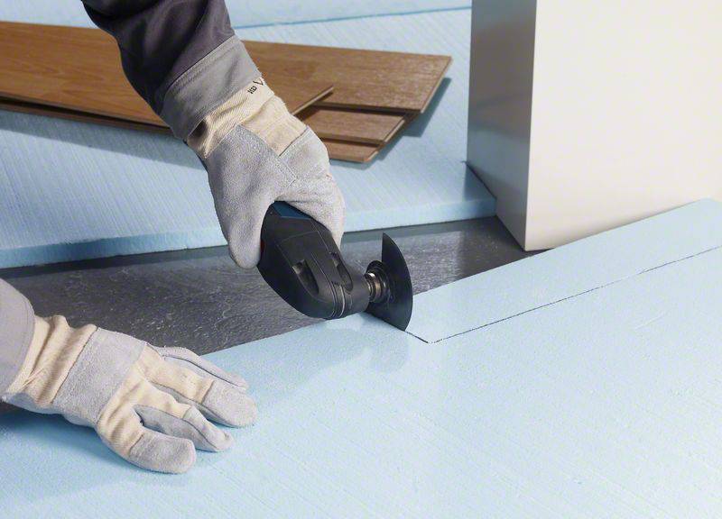 Eine Person mit Arbeitshandschuhen schneidet eine blaue Isolationsplatte mit einem elektrischen Schneidewerkzeug auf Maß.