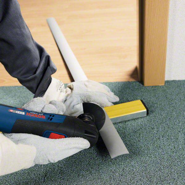 Eine Person benutzt ein Elektrowerkzeug, um einen grauen Rohrabschnitt vor einer hölzernen Tür auf einem grünen Teppich zu schneiden.