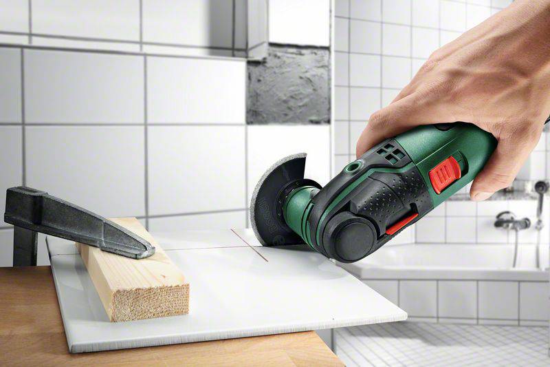 Eine Hand hält ein Elektrowerkzeug und schneidet präzise durch Fliesenmaterial, während ein Metallteil das Werkstück fixiert.