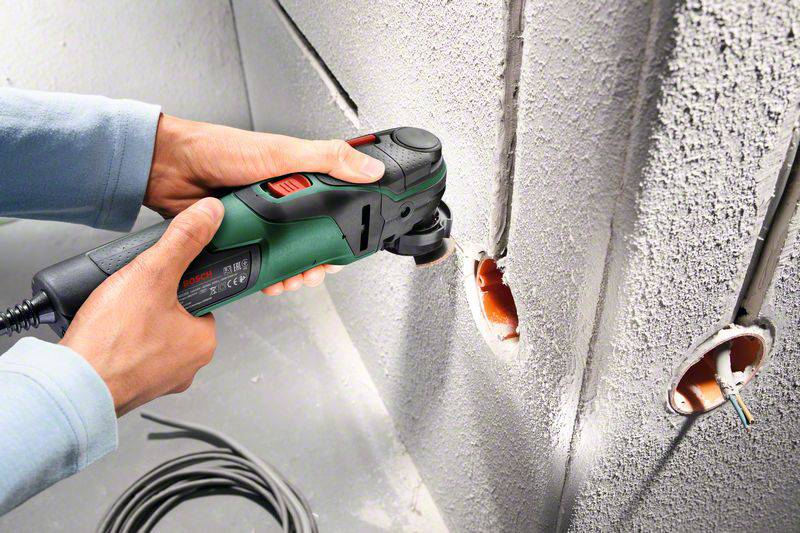 Eine Person verwendet ein elektrisches Multifunktionswerkzeug, um Löcher in eine Betonwand zu bohren. Kabel sind sichtbar.