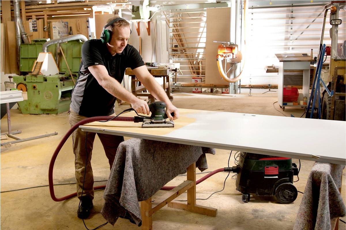 Ein Mann schleift eine große Holzplatte in einer Werkstatt. Er trägt Gehörschutz und konzentriert sich auf die Arbeit.