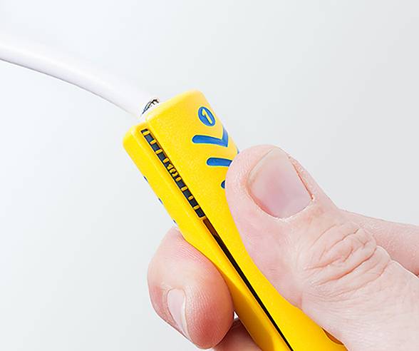 Eine Hand drückt ein gelbes Werkzeug zusammen, um die Isolierung von einem weißen Kabel zu entfernen und den darunter liegenden Metallleiter freizulegen.