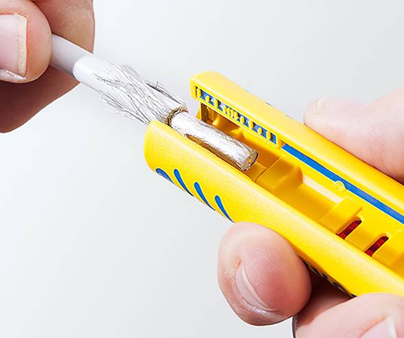 Eine Nahaufnahme von Händen, die einen gelben Abisolierwerkzeug verwenden, um die Isolierung von einem Kabel zu entfernen und dabei die inneren Metallstränge freizulegen.