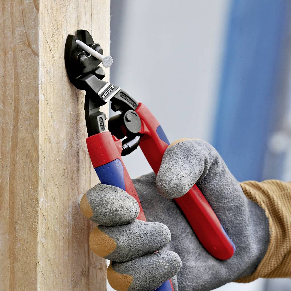 Eine Person in Arbeitshandschuhen schneidet mit einer roten Zange einen Nagel aus einem Holzbrett.